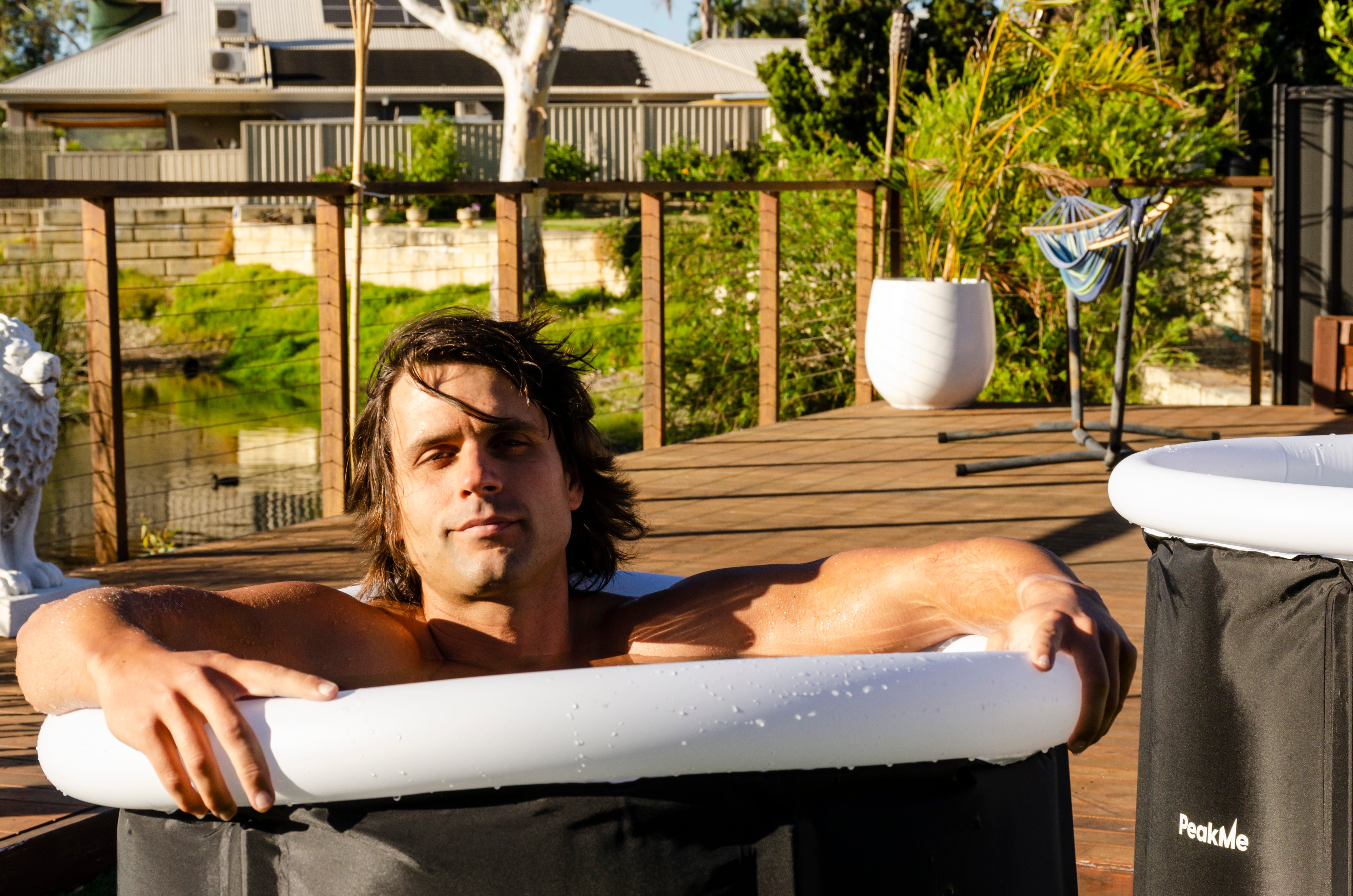 Relaxed man sitting in a PeakMe portable ice bath, enjoying the invigorating effects of cold therapy in a sun-drenched, peaceful backyard retreat.