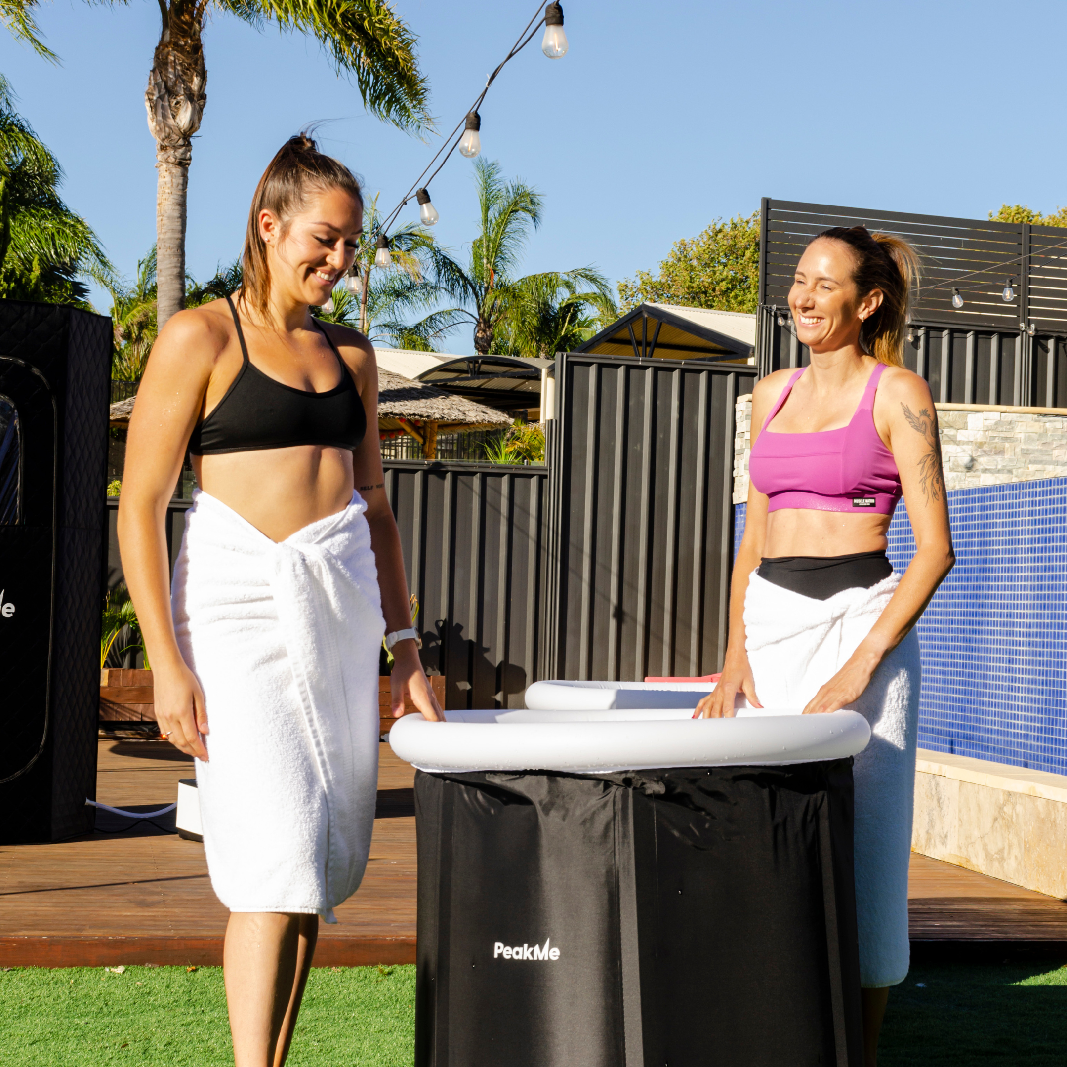 Two smiling women in activewear next to a portable PeakMe ice bath, preparing for cold therapy in an outdoor setting, promoting recovery and wellness.