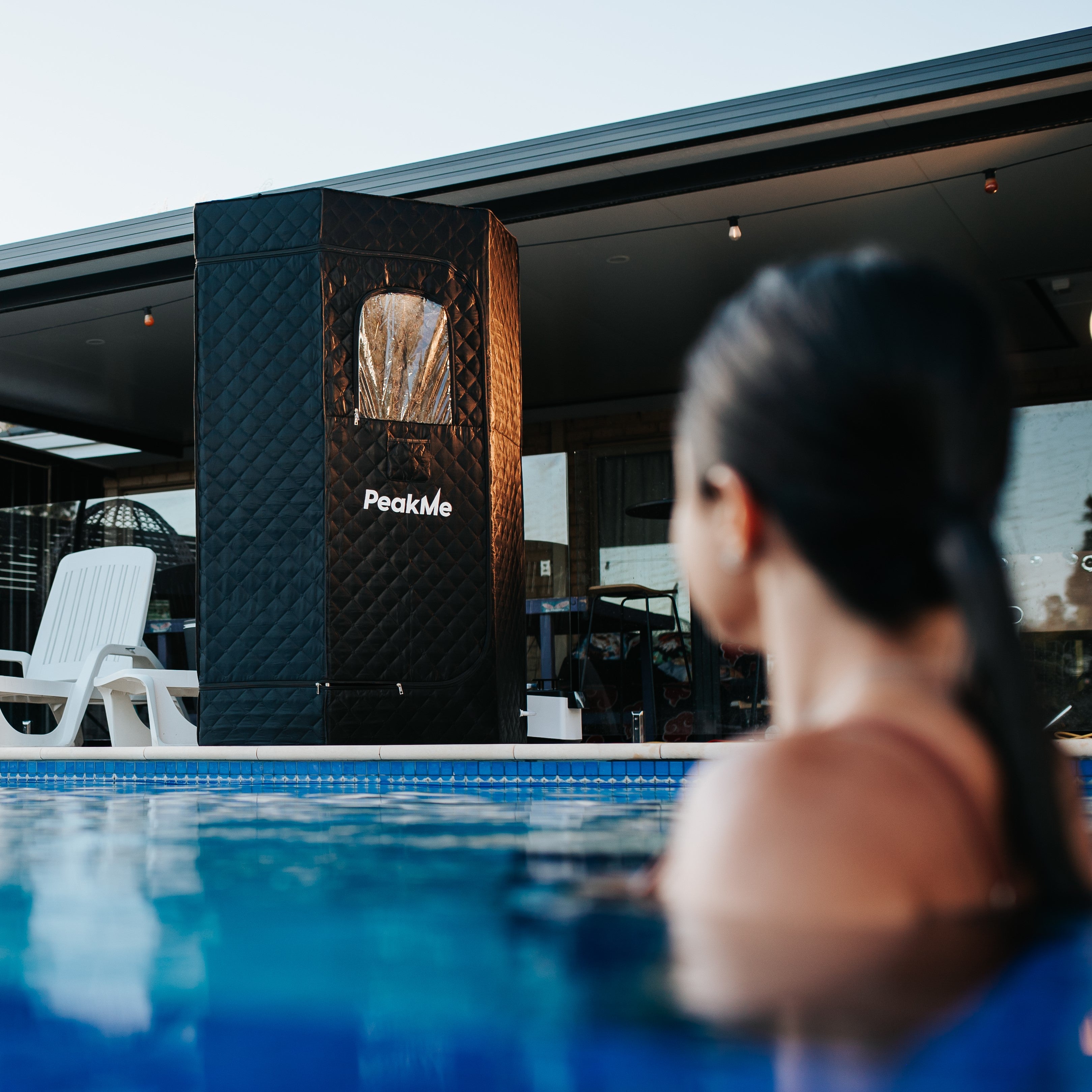 Peaceful poolside setting with a woman looking towards the PeakMe Retreat Steam Sauna, highlighting luxury wellness in a home environment with comfortable outdoor seating.