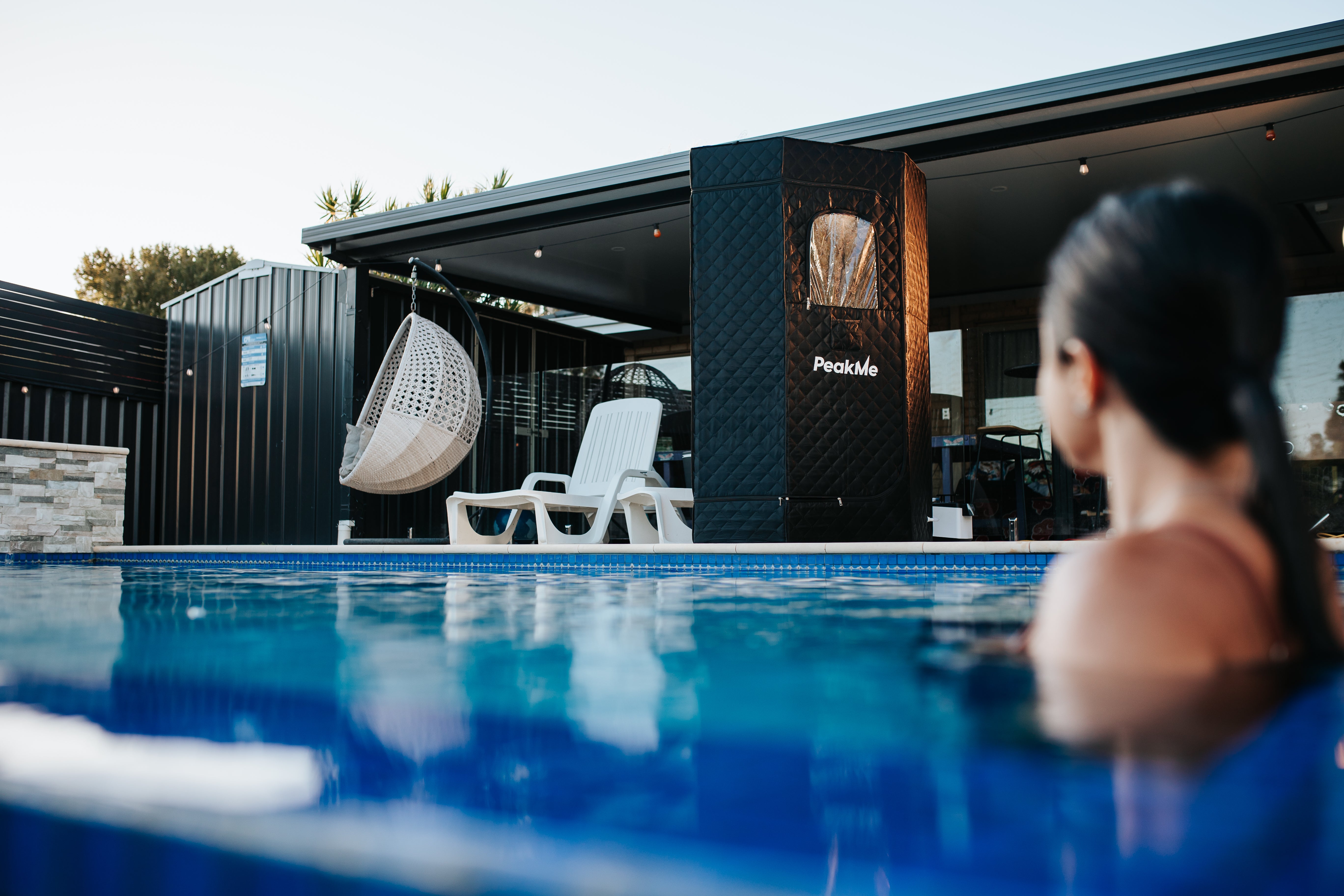 Peaceful poolside setting with a woman looking towards the PeakMe Retreat Steam Sauna, highlighting luxury wellness in a home environment with comfortable outdoor seating.