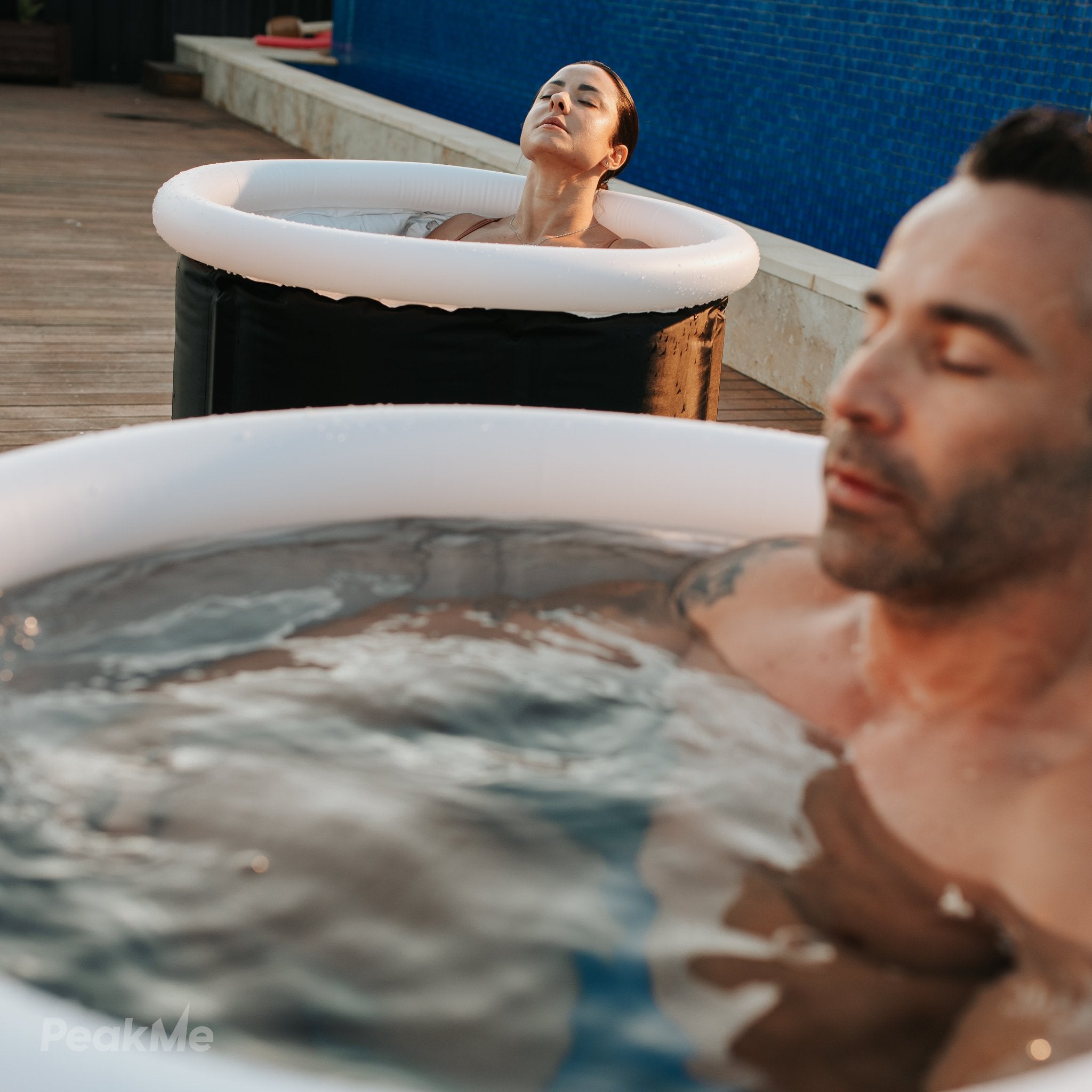 Man and woman in personal PeakMe Focus ice baths relaxing outdoors with the PeakMe logo visible, experiencing the benefits of cold immersion therapy at home.